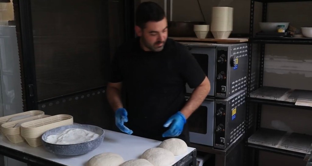 Baker in a black shirt and blue gloves working at a bakery shaping bench with dough portions, oval bannetons and two deck ovens