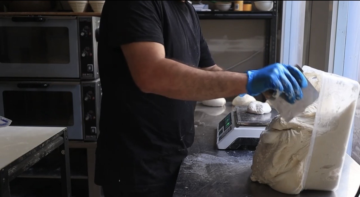 A baker dividing sourdough dough with a metal bench scraper beside a digital scale