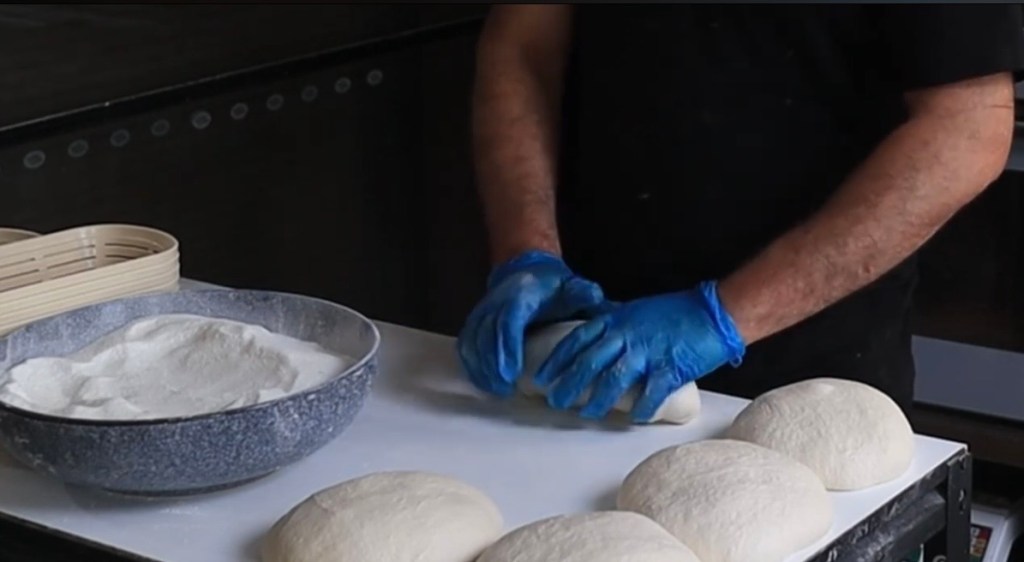 Close-up of a baker's blue-gloved hands gently shaping sourdough dough into a loaf on a bakery worktable